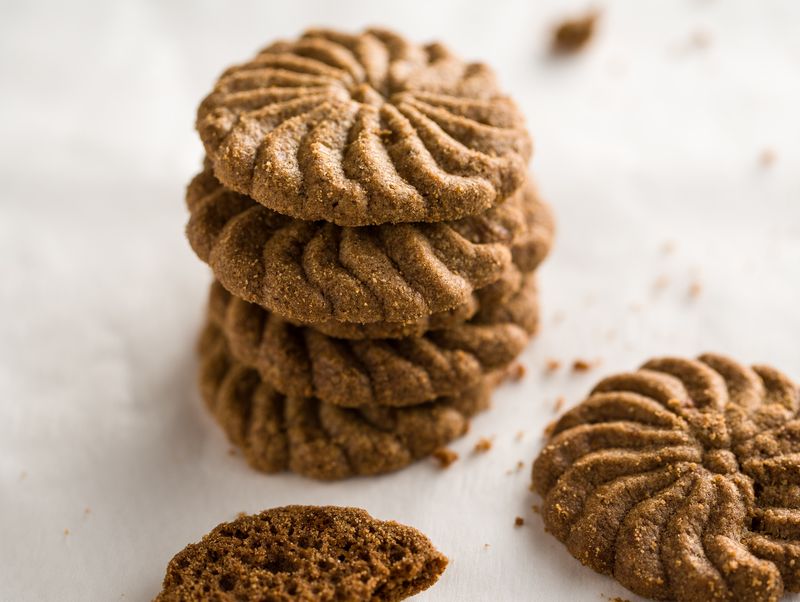Gestapelte, spiralförmig geprägte braune Kekse auf hellem Untergrund, mit verstreuten Krümeln und einem gebrochenen Keks im Vordergrund.