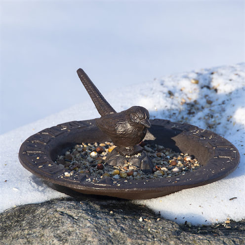 Een gietijzeren zonnewijzer in de vorm van een vogelbad met een vogelfiguur op de rand, staande in de sneeuw.