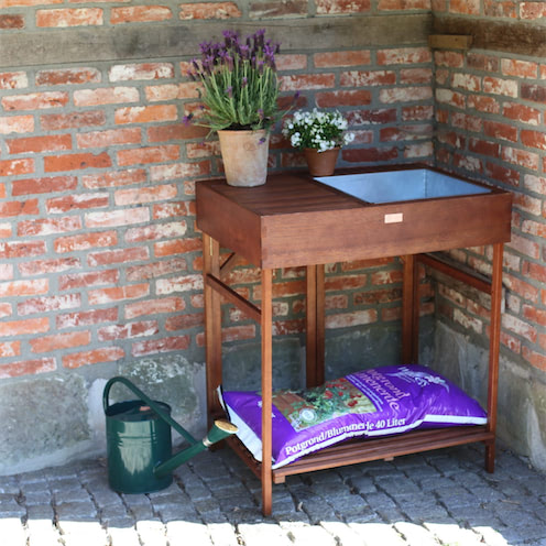 Een hardhouten oppottafel met een zinken bak en een opbergplank eronder. Op de tafel staan bloempotten en een gieter staat ernaast.