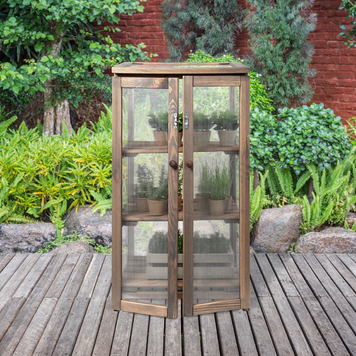 Een houten kas met glazen panelen en deuren, gevuld met verschillende planten in potten, staat op een houten terras.