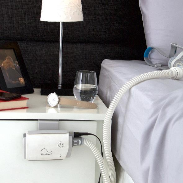 ResMed AirMini CPAP-Reisegerät mit Betthalterung am Nachttisch befestigt, neben dem Bett mit angeschlossenem Atemschlauch.