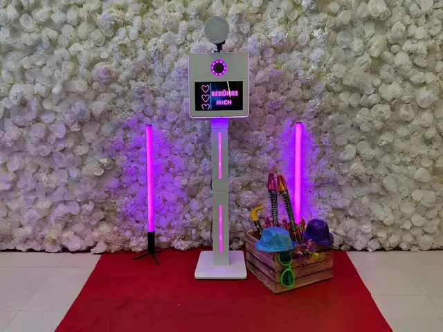 Fotobox auf einer Hochzeit mit LED-Lichtern, Requisitenkiste und Blumenwand – moderne Selfiebox mit Touchscreen.