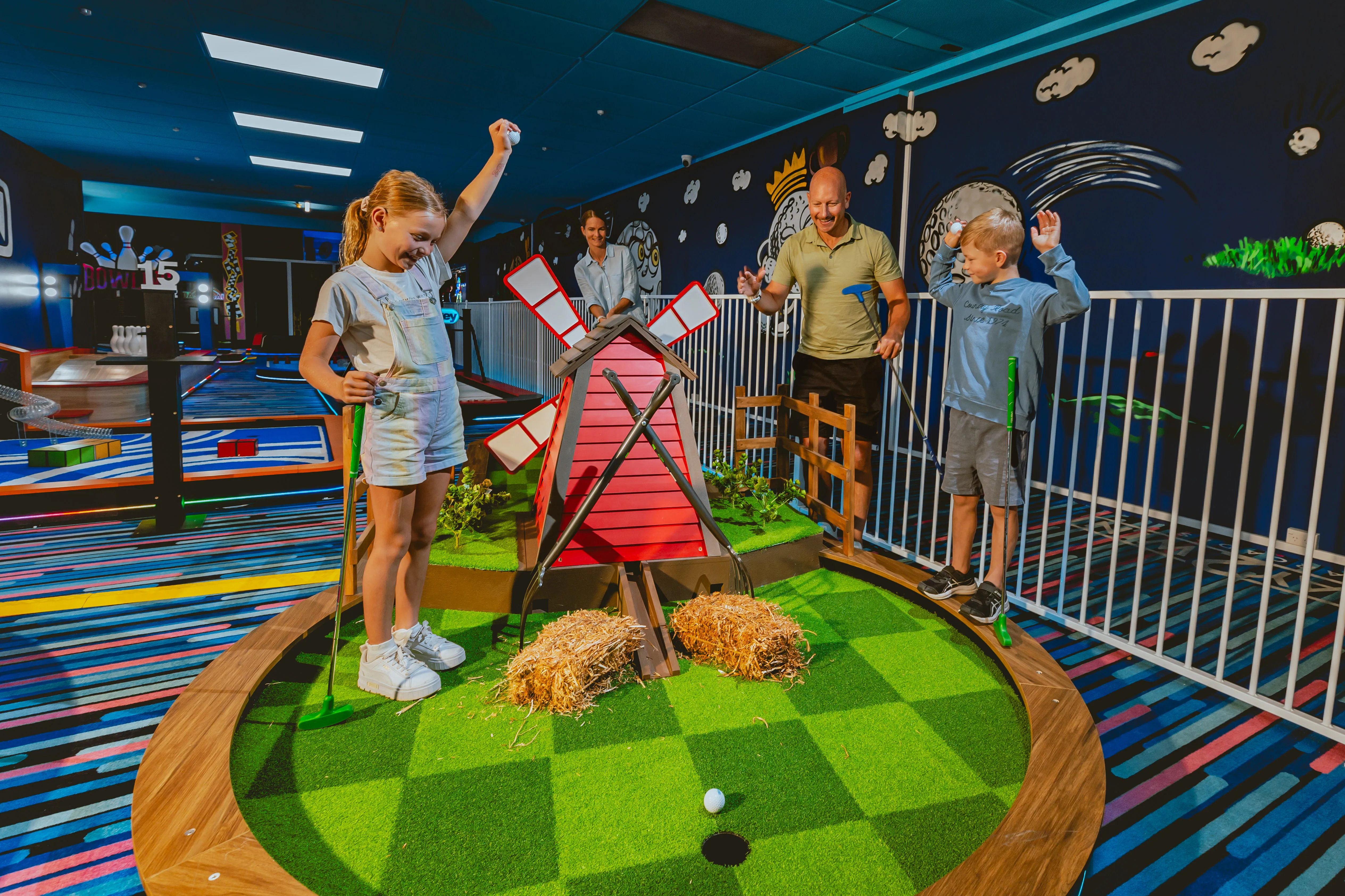 Kinder und ein Erwachsener jubeln auf einer farbenfrohen Indoor-Minigolfbahn mit Windmühlen-Hindernis, umgeben von verspielter Wandgestaltung und buntem Boden.