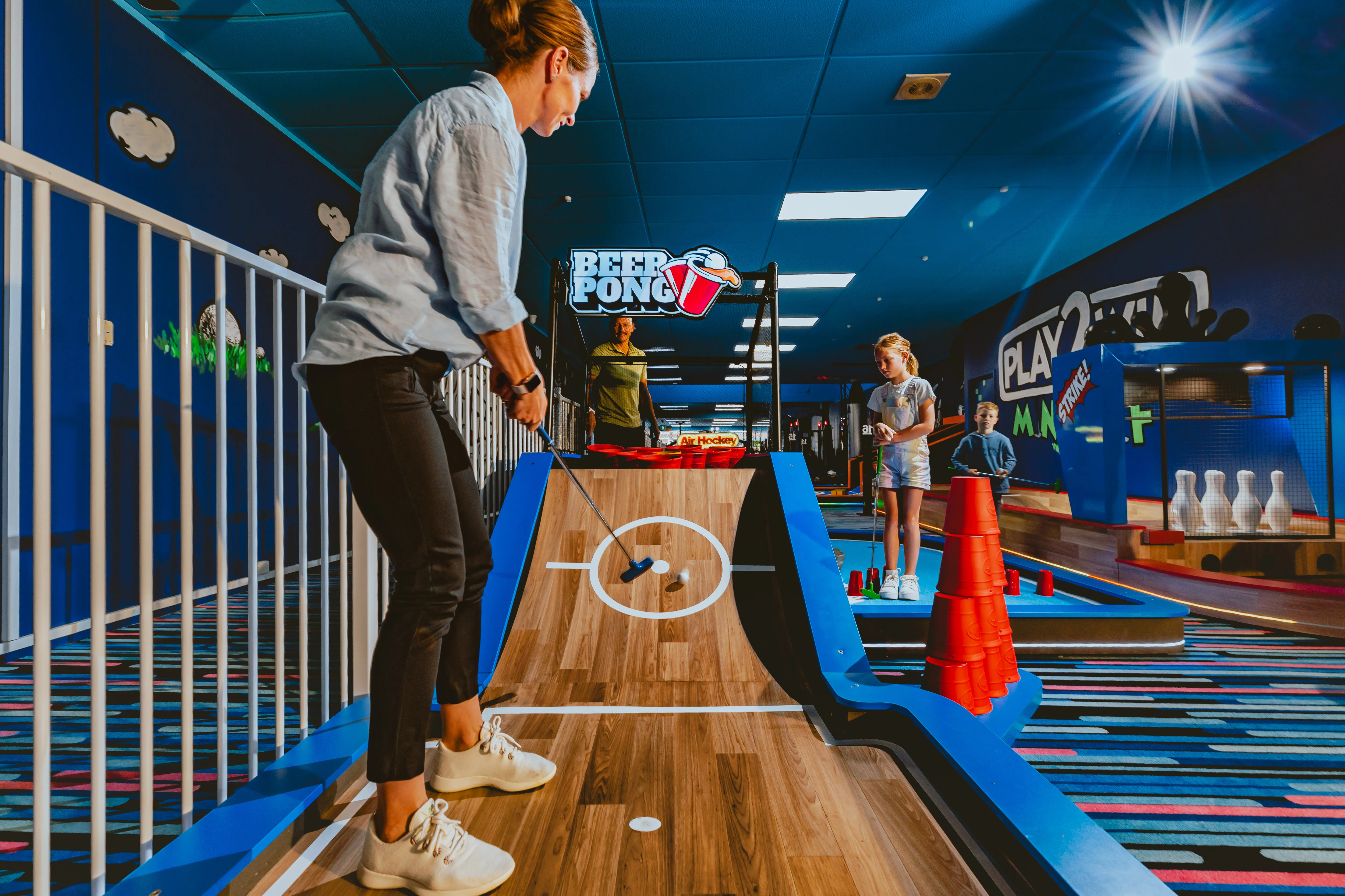 Erwachsene Person spielt Abenteuer Minigolf auf einer thematisch gestalteten Beer-Pong-Bahn bei Laserstar® Oldenburg. Kinder und weitere Spielstationen im Hintergrund in farbenfroher Indoor-Freizeitumgebung.