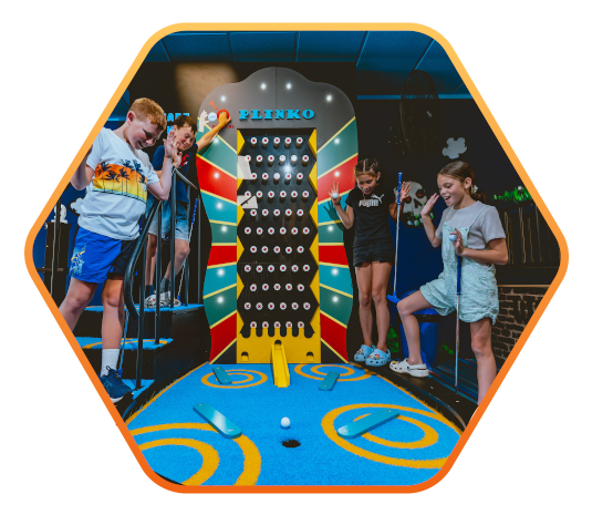 Vier Kinder spielen begeistert an einem bunten „Plinko“-Spielautomaten in einer Indoor-Spielhalle. Eine Kugel rollt über ein leuchtend blaues Spielfeld mit gelben Markierungen. Die Szene ist in einem sechseckigen Rahmen mit orangefarbener Umrandung darges