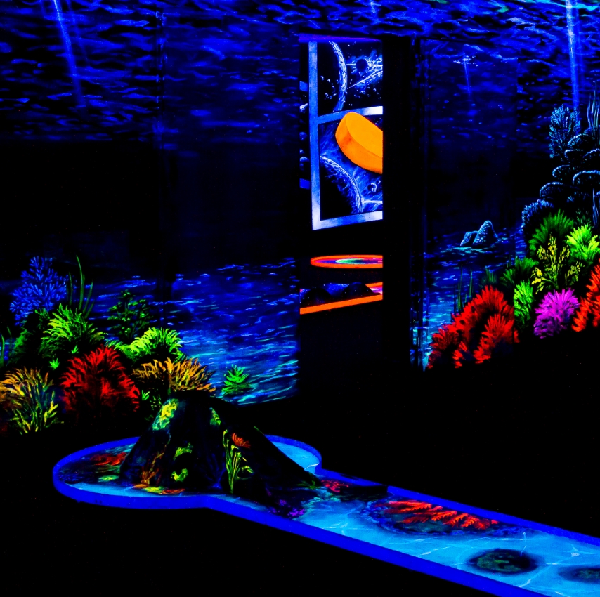 Eine leuchtende Schwarzlicht-Minigolfbahn in der SIMIGOLF-Anlage Asperg mit einem fantasievollen Mix aus Unterwasserwelt und futuristischem Raumthema. Im Vordergrund erhebt sich ein kleiner Vulkan mit bunten Korallen drumherum, der als Hindernis auf der B