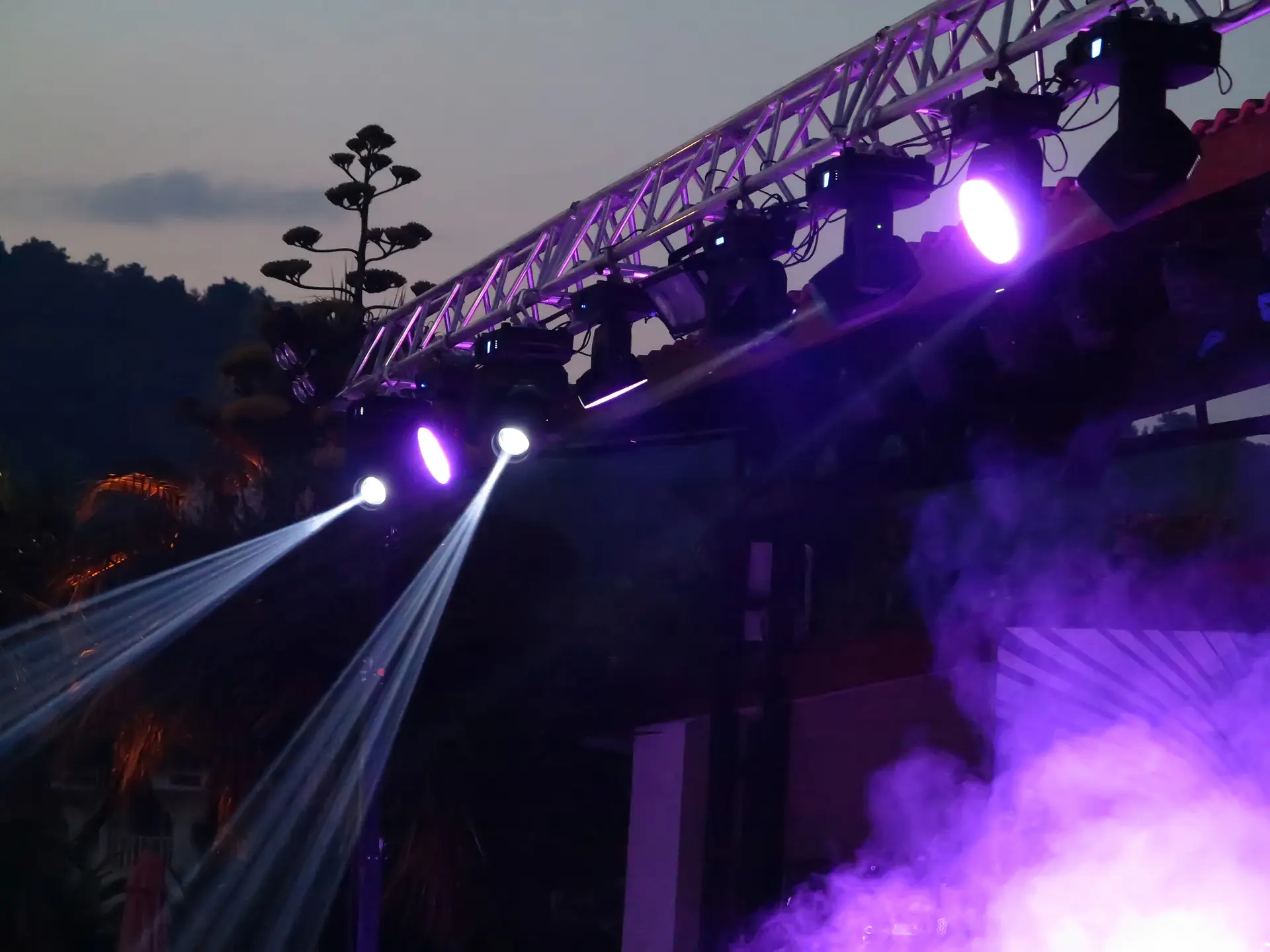 Open-Air Lichtshow mit Moving-Heads, Nebel und LED-Scheinwerfern an einer Traverse bei Abenddämmerung.