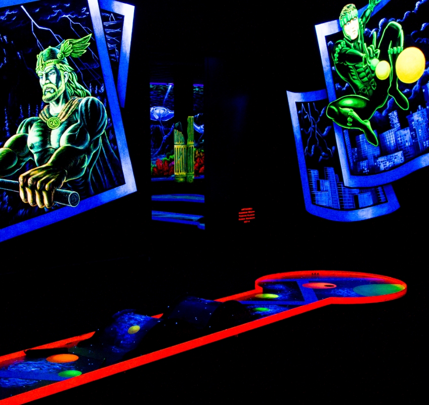 Eine actionreiche Schwarzlicht-Minigolfbahn aus der SIMIGOLF-Anlage Asperg, inspiriert vom Superhelden- und Comic-Universum. Links ist ein mächtiger Held mit Flügelhelm und Hammer dargestellt, während rechts ein futuristischer Kämpfer mit leuchtenden Kuge