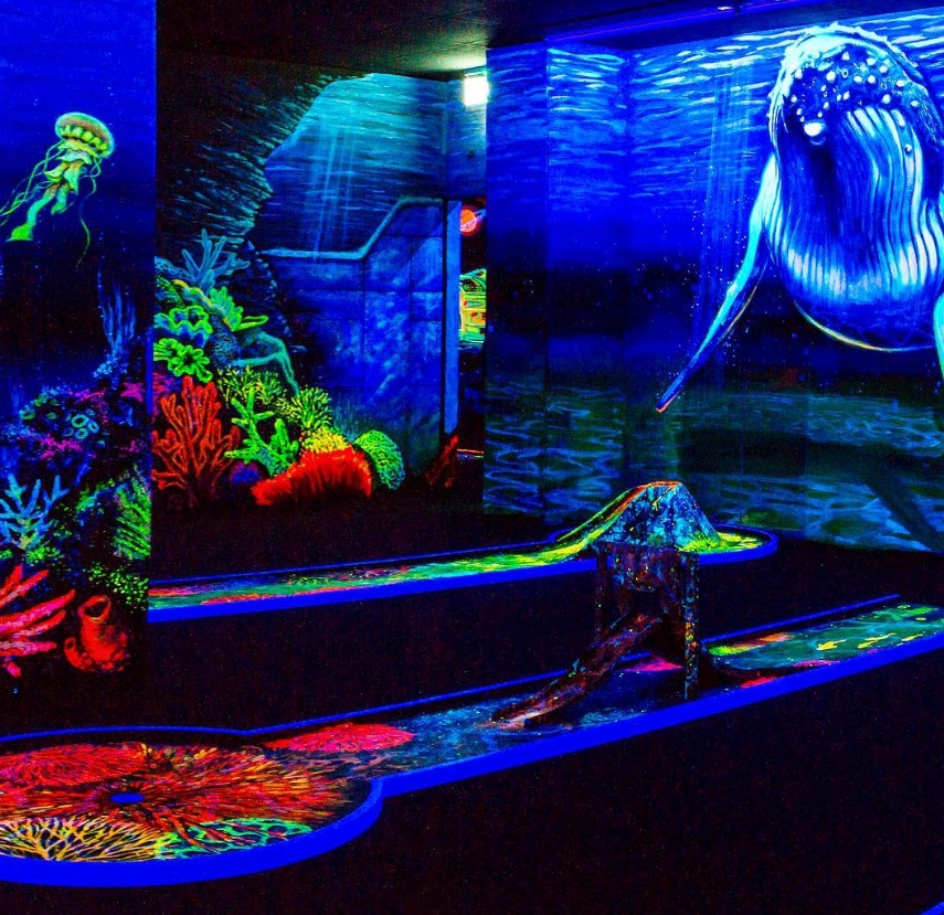 Eine beeindruckende Schwarzlicht-Minigolfbahn mit einem atemberaubenden Unterwasserdesign. Ein riesiger, leuchtend blauer Wal schwimmt majestätisch durch die Szene, während bunte Korallen und eine geheimnisvolle Meereslandschaft die Umgebung prägen. Links