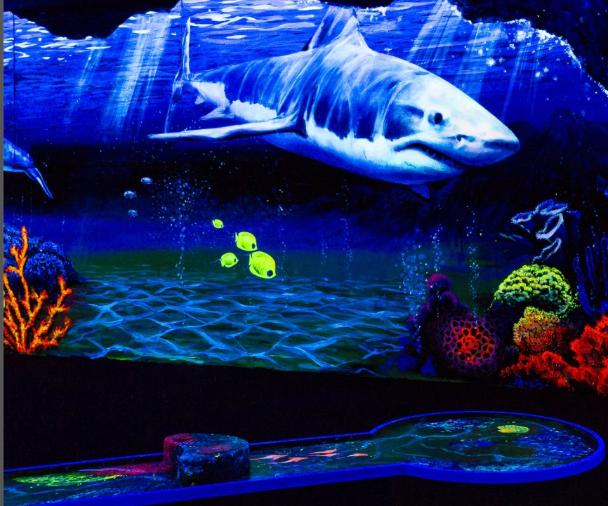 Eine faszinierende Schwarzlicht-Minigolfbahn mit einem leuchtenden Unterwasser-Motiv. Ein imposanter weiß-blauer Hai schwimmt durch die Szene, umgeben von bunten Korallen und leuchtenden Fischen. Die Minigolfbahn im Vordergrund reflektiert das Schwarzlich