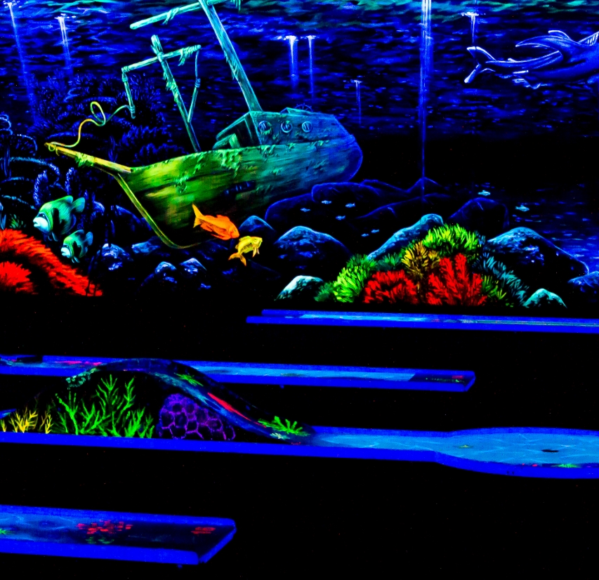 Eine atmosphärische Schwarzlicht-Minigolfbahn in der SIMIGOLF-Anlage Asperg mit einer eindrucksvollen Unterwasserszenerie. Im Hintergrund liegt ein gesunkenes, grünlich leuchtendes Schiffswrack zwischen Felsen und farbenfrohen Korallen. Verschiedene Fisch