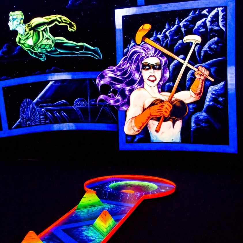 Eine energiegeladene Schwarzlicht-Minigolfbahn in der SIMIGOLF-Anlage Asperg mit einem knalligen Comic-Design. Im Mittelpunkt steht eine gezeichnete Superheldin mit violettem Haar, Sonnenbrille und zwei Minigolf-Schlägern in Kampfpose. Im Hintergrund schw