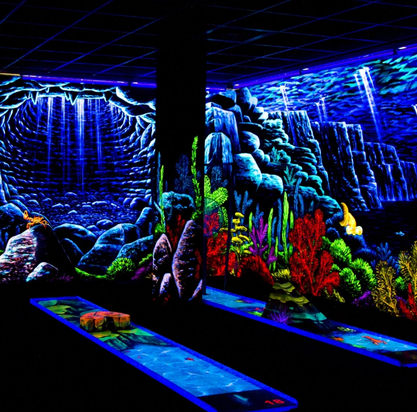 Eine faszinierende Schwarzlicht-Minigolfbahn in der SIMIGOLF-Anlage Asperg mit detailreicher Höhlen- und Unterwasserlandschaft. Leuchtende Felsen, bunte Korallen und ein mystischer Höhleneingang mit herabfallendem Lichtstrahl bilden die Kulisse. Im Vorder