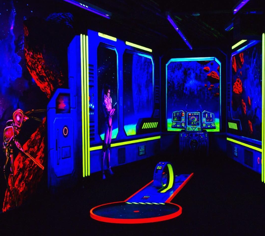 Eine faszinierende Schwarzlicht-Minigolfbahn mit einem futuristischen Weltraumthema. Die Szene zeigt eine Raumstation mit leuchtenden blauen Fenstern, durch die ein Blick ins All mit roten Asteroiden möglich ist. Eine humanoide Figur in einem High-Tech-An