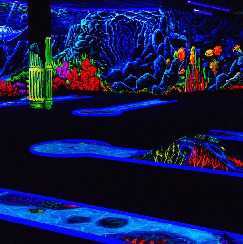 Eine farbenprächtige Schwarzlicht-Minigolfanlage der SIMIGOLF-Location in Asperg. Die Szene zeigt mehrere fluoreszierende Minigolfbahnen, eingebettet in eine fantasievolle Unterwasserwelt. Im Hintergrund befinden sich bunte Korallen, leuchtende Fische und