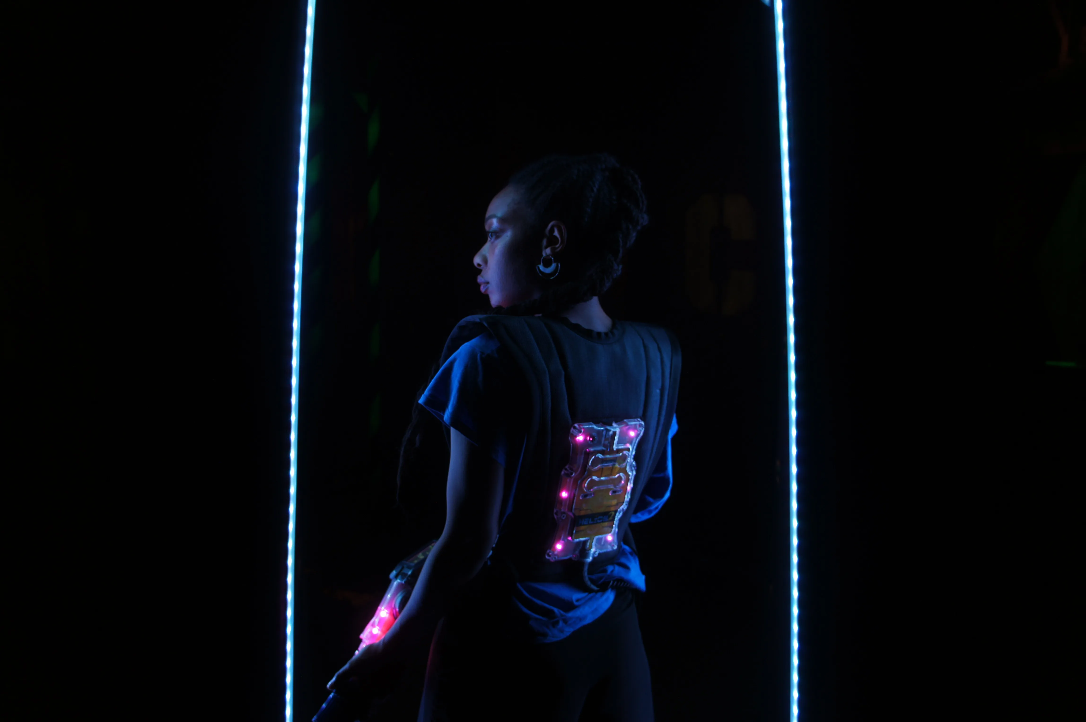 Eine junge Frau steht mit einer Lasertag Weste in einem Dunklen Raum, mit dem Rüücken zur Kamera, Blick nach links gewendet. Links und rechts neben ihr zwei Neonröhren.