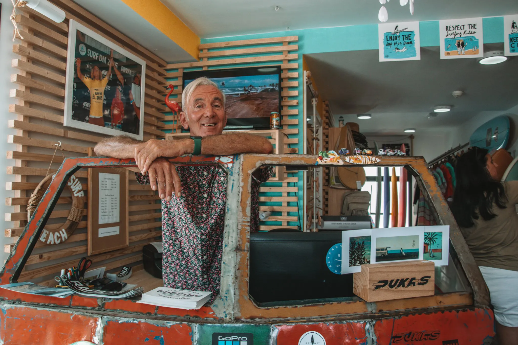 Ein älterer Herr steht in einem Surf-Shop und lehnt sich freundlich über die Theke und lächelt in die Kamera. 