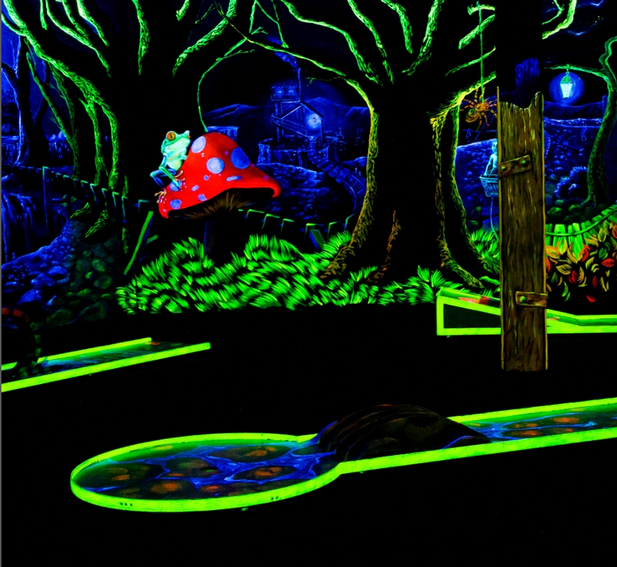 Eine fantasievolle Schwarzlicht-Minigolfbahn mit märchenhaftem Waldmotiv in der SIMIGOLF-Anlage Asperg. Im Zentrum sitzt ein großer, leuchtender Frosch mit Krone auf einem rot-weißen Fliegenpilz. Umgeben von leuchtend grünen Gräsern, verwunschenen Bäumen 