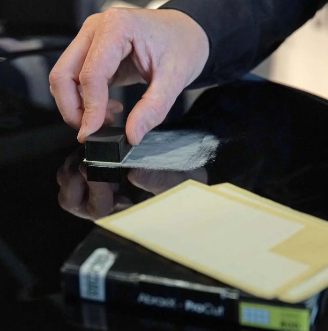 Eine Hand trägt mit einem kleinen schwarzen Schaumstoffblock eine weiße Polierpaste auf eine glänzende schwarze Autooberfläche auf, im Vordergrund ist eine Schachtel Abraz-ProCut-Polierpads zu sehen.