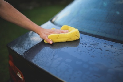 Die Hand einer Person wäscht mit einem gelben Schwamm den nassen, dunklen Kofferraum eines Autos, wobei Wassertropfen auf der Oberfläche sichtbar sind.