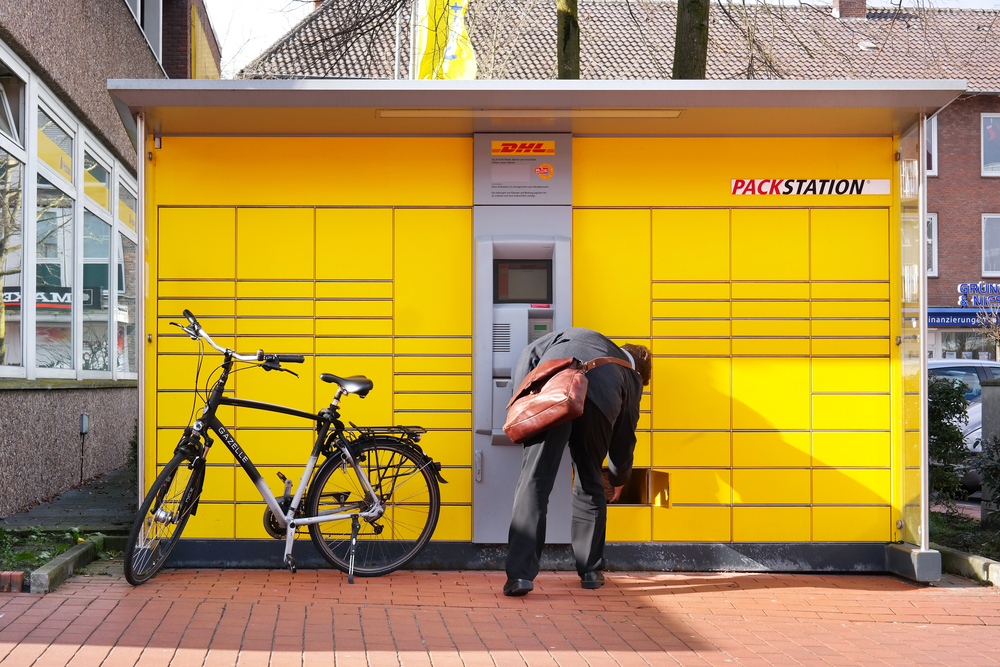 Text: Auf einem Grundstück steht eine Packstation, Mann im grauen Anzug und Fahrrad, Paket zur Abholung.