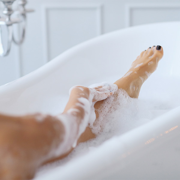 Text: Weißer Hintergrund, zwei Beine in einer Badewanne, bedeckt mit Badeschaum.