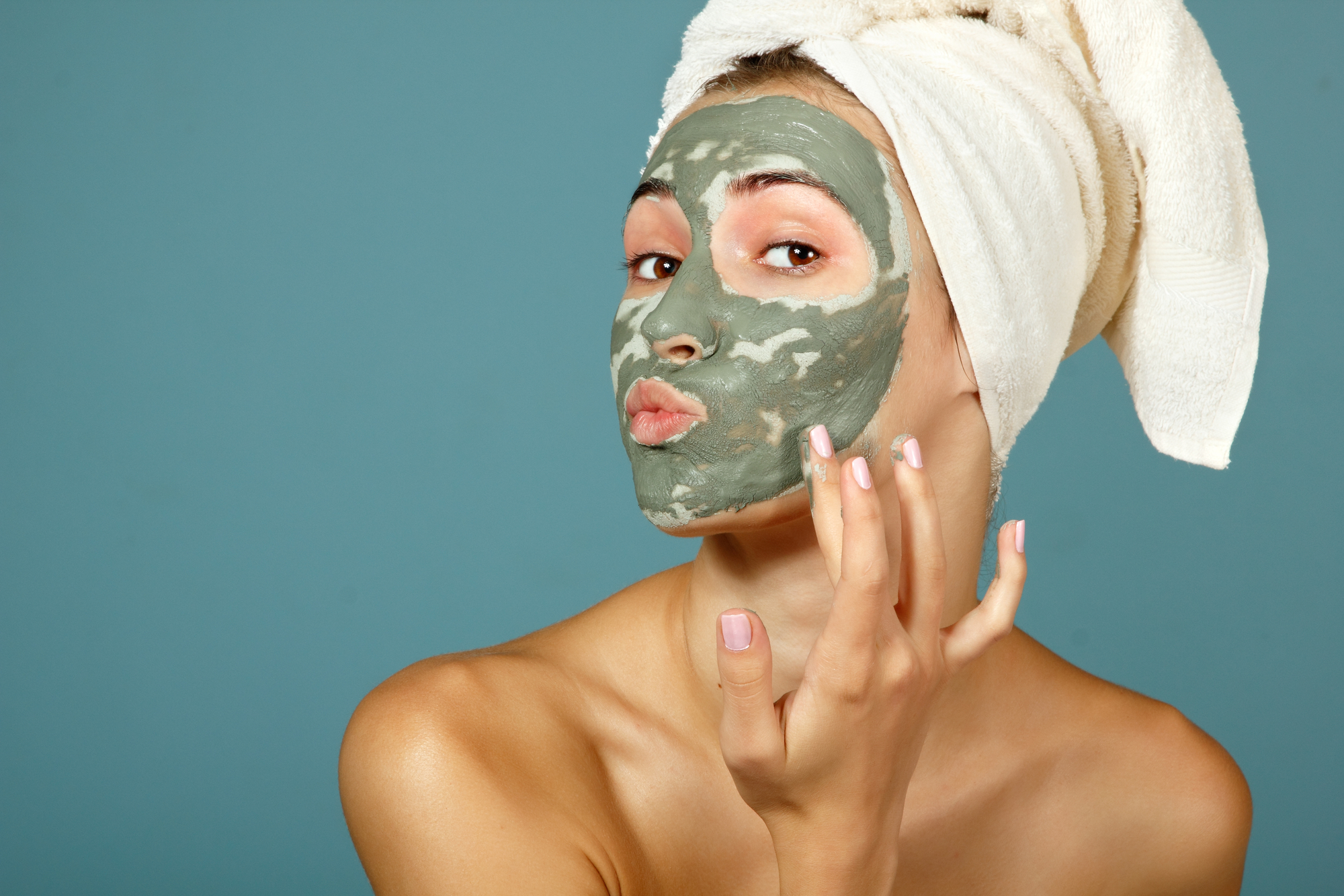 Text: Hintergrund Farbe petrol, eine Dame mit einer grünen Gesichtsmaske, bedeckt mit einem Handtuch auf dem Kopf.