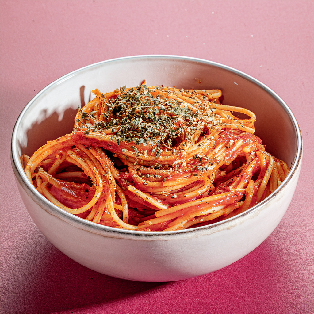 Nahaufnahme von Spaghetti mit Tomatensauce, bestreut mit getrockneten Kräutern, serviert in einer weißen Keramikschüssel auf rosa Untergrund.