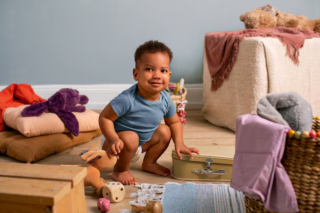 Ein Kleinkind in einem blauen T-Shirt steht auf dem Boden und hebt eine kleine Kiste an. Um es herum sind Spielsachen, Decken und ein Korb mit weiteren Textilien.