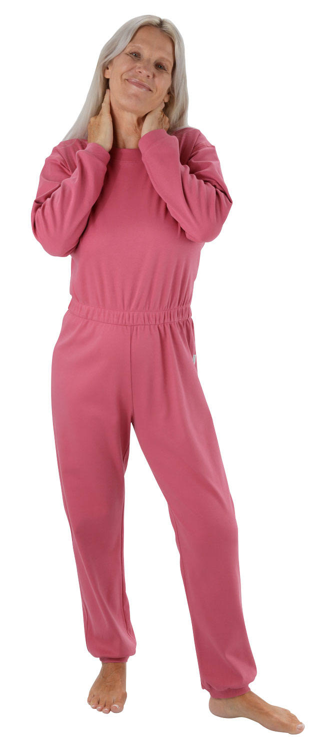 Eine ältere Frau trägt einen pinken Jumpsuit. Sie steht entspannt da, berührt ihren Hals und lächelt. Der Hintergrund ist neutral und hebt die Kleidung hervor.