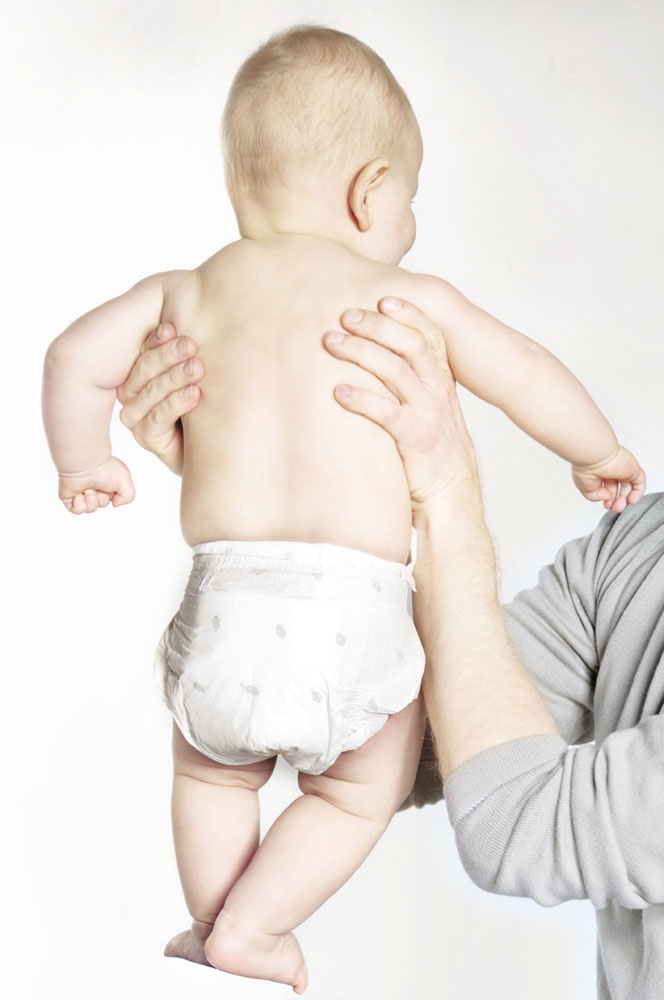 Ein Baby wird von einer Person mit einer Hand gehalten. Das Baby trägt eine windelformige Bekleidung. Der Hintergrund ist ein blasses Weiß, ohne ablenkende Elemente.
