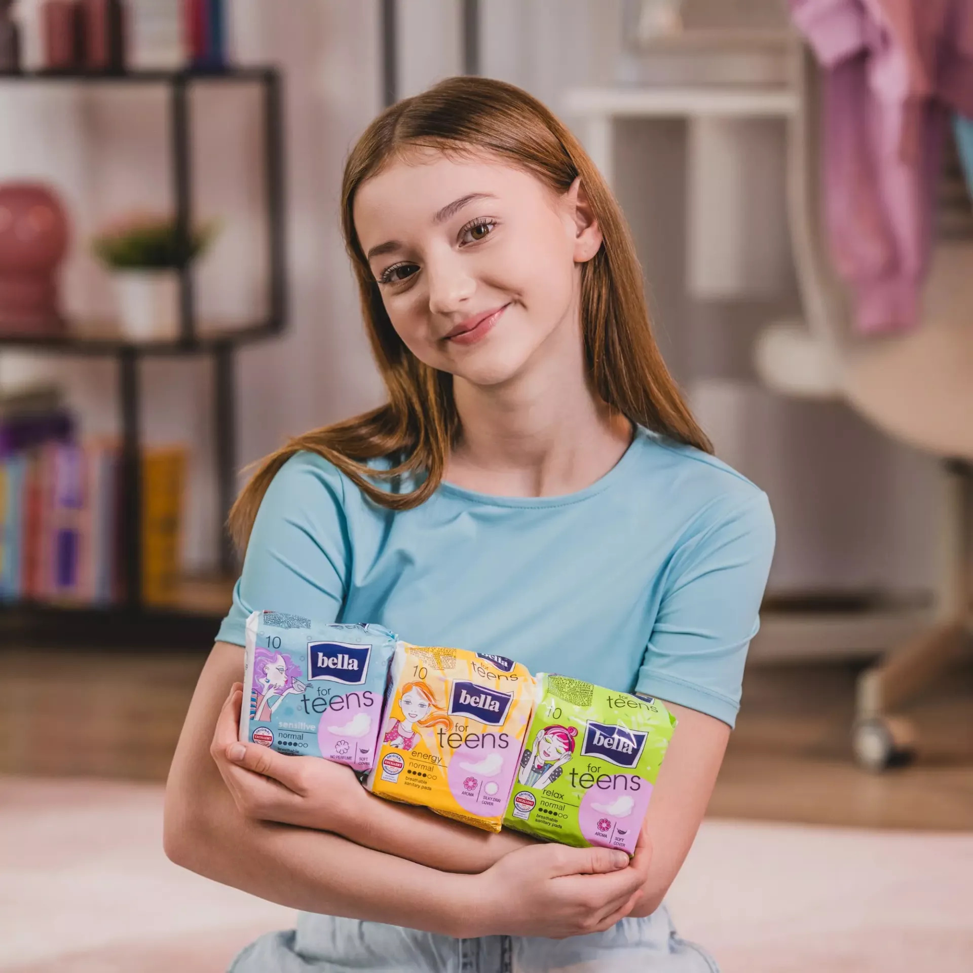 Ein Mädchen in einem hellblauen Shirt hält drei Packungen Bella Produkte für Teenager. Im Hintergrund sind Bücherregale und ein Stuhl sichtbar. Die Packungen sind farbenfroh und mit Zeichnungen.