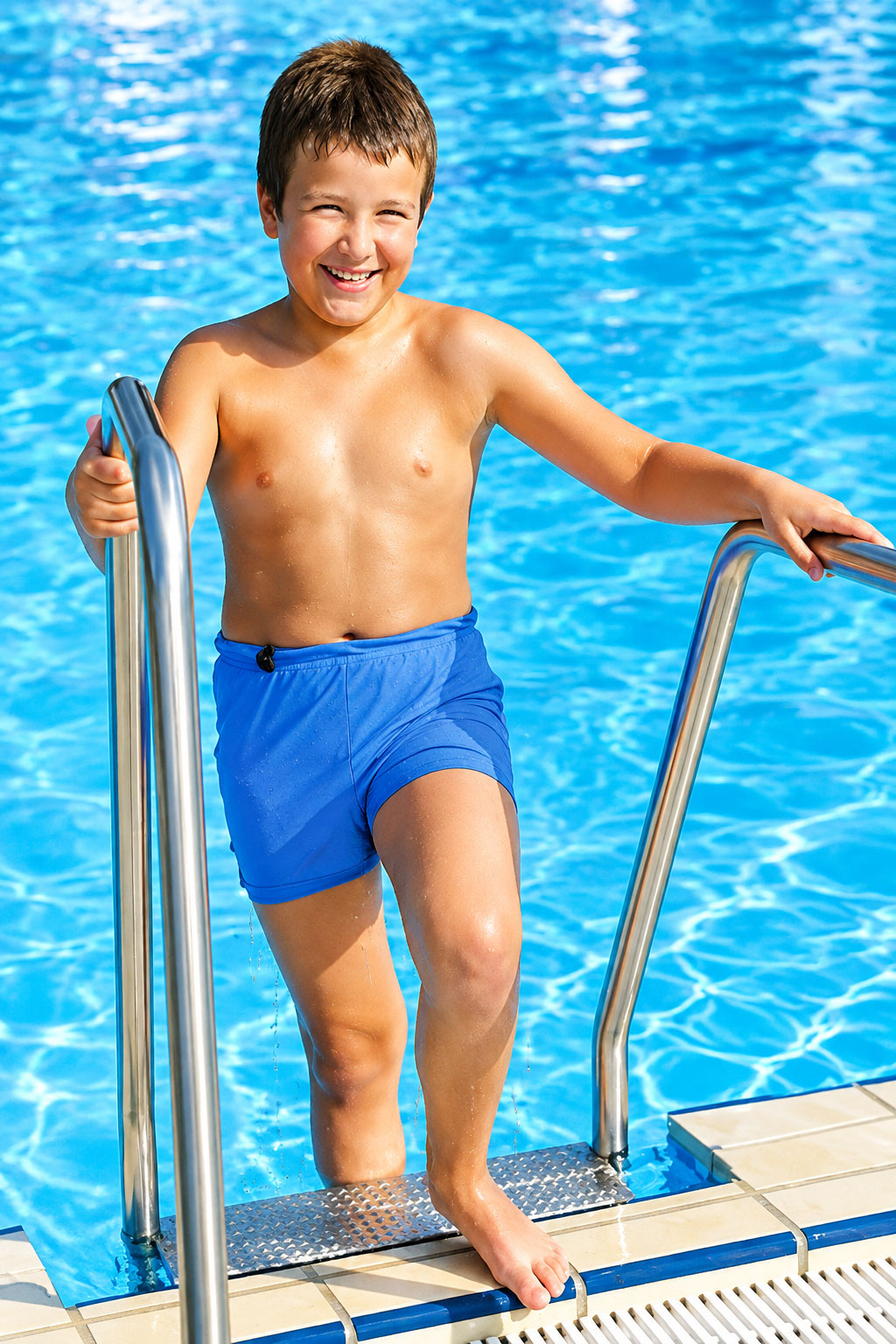 Ein Junge mit einem breiten Lächeln steht auf einer Schwimmbadleiter und blickt nach vorne. Er trägt blaue Schwimmshorts und hat nasses Haar. Im Hintergrund sieht man glitzerndes Wasser.