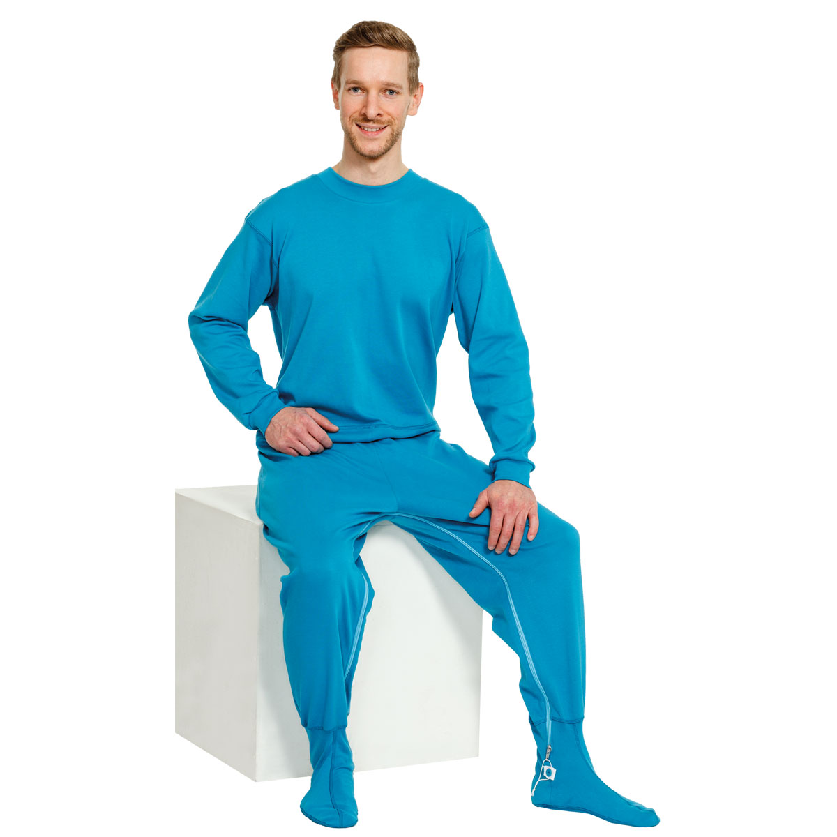 Mann sitzt auf einer weißen Box, trägt einen einfarbigen, hellblauen Langarmanzug mit passenden Socken. Er lächelt und präsentiert die bequeme Kleidung in einem neutralen Umfeld.
