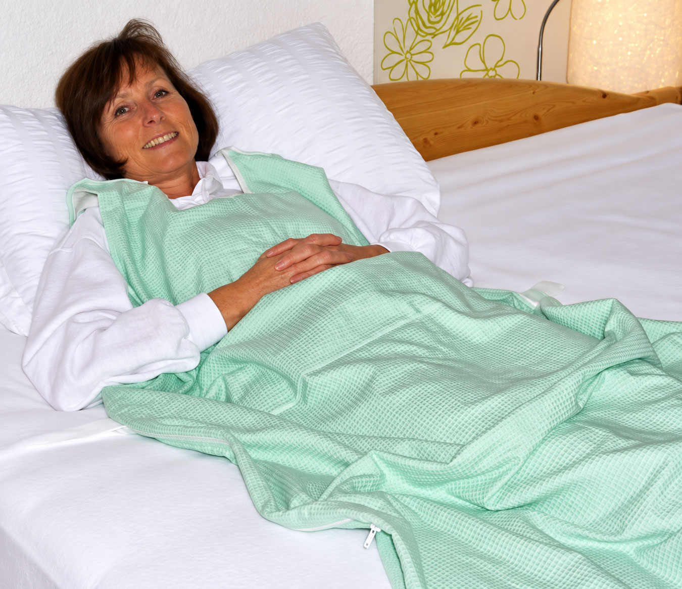 Eine Frau liegt entspannt auf einem Bett, trägt eine mintgrüne Pflegebekleidung und hat die Hände auf dem Bauch gefaltet. Der Hintergrund zeigt eine helle Wand und eine Lampe.