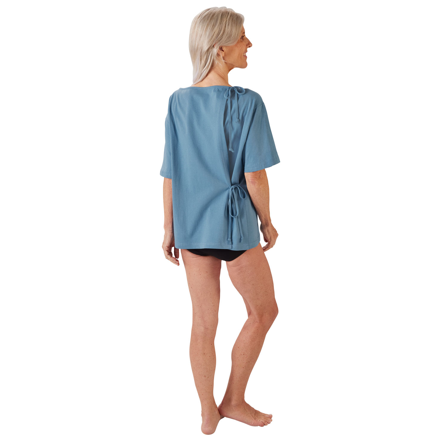 Eine ältere Frau steht mit dem Rücken zur Kamera, trägt ein hellblaues Shirt mit kurzen Ärmeln, das hinten zusammengebunden ist. Sie hat schwarze Unterwäsche und zeigt lächelnd ihre Beine.