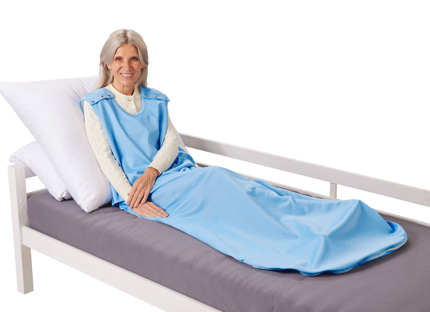 Eine lächelnde ältere Frau sitzt auf einem Bett mit grauem Bezug. Sie trägt einen blauen Überwurf und hat eine helle Bluse an. Sie ist von Kopfkissen umgeben.