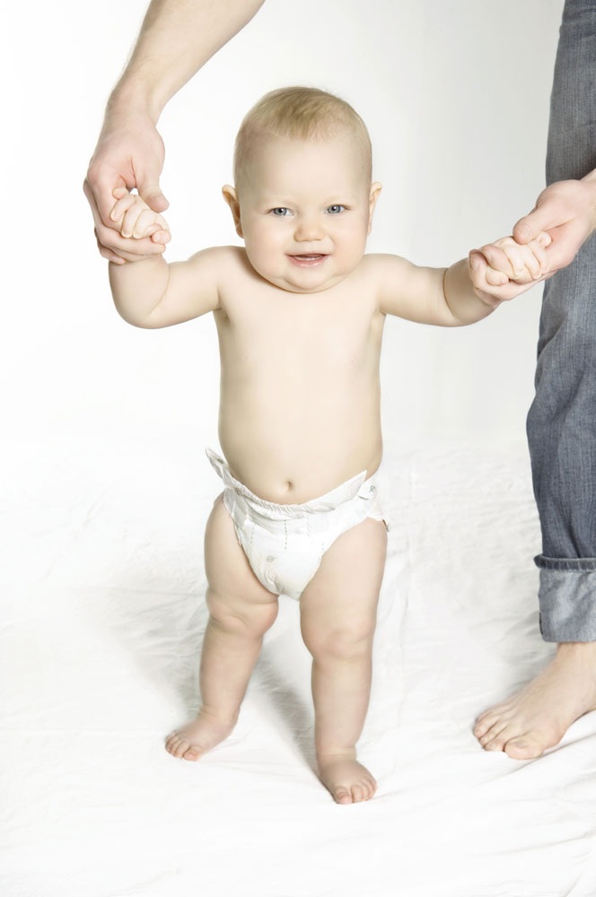 Ein Baby steht in einer Windel und hält mit beiden Händen die Hände eines Erwachsenen. Der Hintergrund ist weiß und schlicht, was den Fokus auf das Baby lenkt.