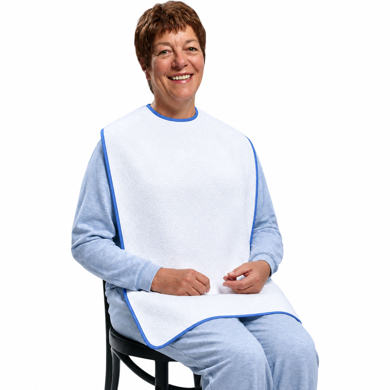 Eine sitzende Frau trägt ein großes, weißes Lätzchen mit blauen Rändern. Sie lächelt freundlich, während sie ihre Hände auf ihrem Schoß liegen hat. Der Hintergrund ist weiß und neutral.