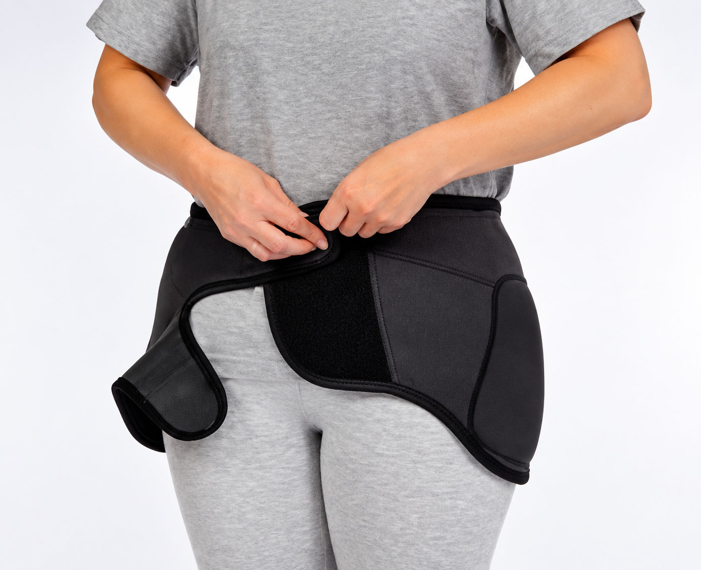Eine Person trägt eine schwarze Inkontinenzhose und befestigt den Verschluss. Sie steht vor einem hellen Hintergrund und trägt ein graues T-Shirt sowie graue leggings.