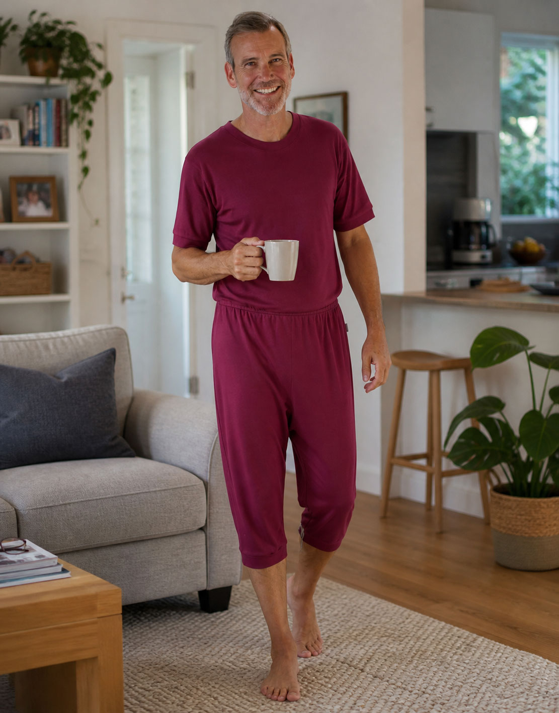 Ein Mann trägt einen burgunderfarbenen Schlafanzug mit kurzen Hosen und lächelt, während er mit einer Tasse in der Hand durch einen hellen Wohnraum läuft. Hintergrund ist eine Couch und eine Pflanze.
