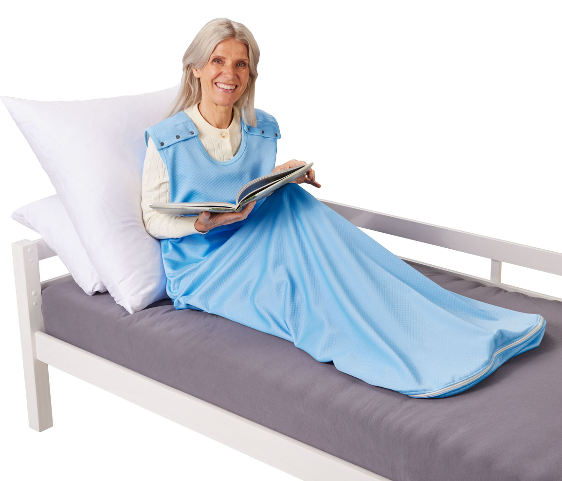 Eine ältere Frau sitzt im Bett, trägt eine blaue Decke mit Armöffnungen und liest ein Buch. Sie lächelt, während sie auf einem grauen Bettlaken und Kissen sitzt.