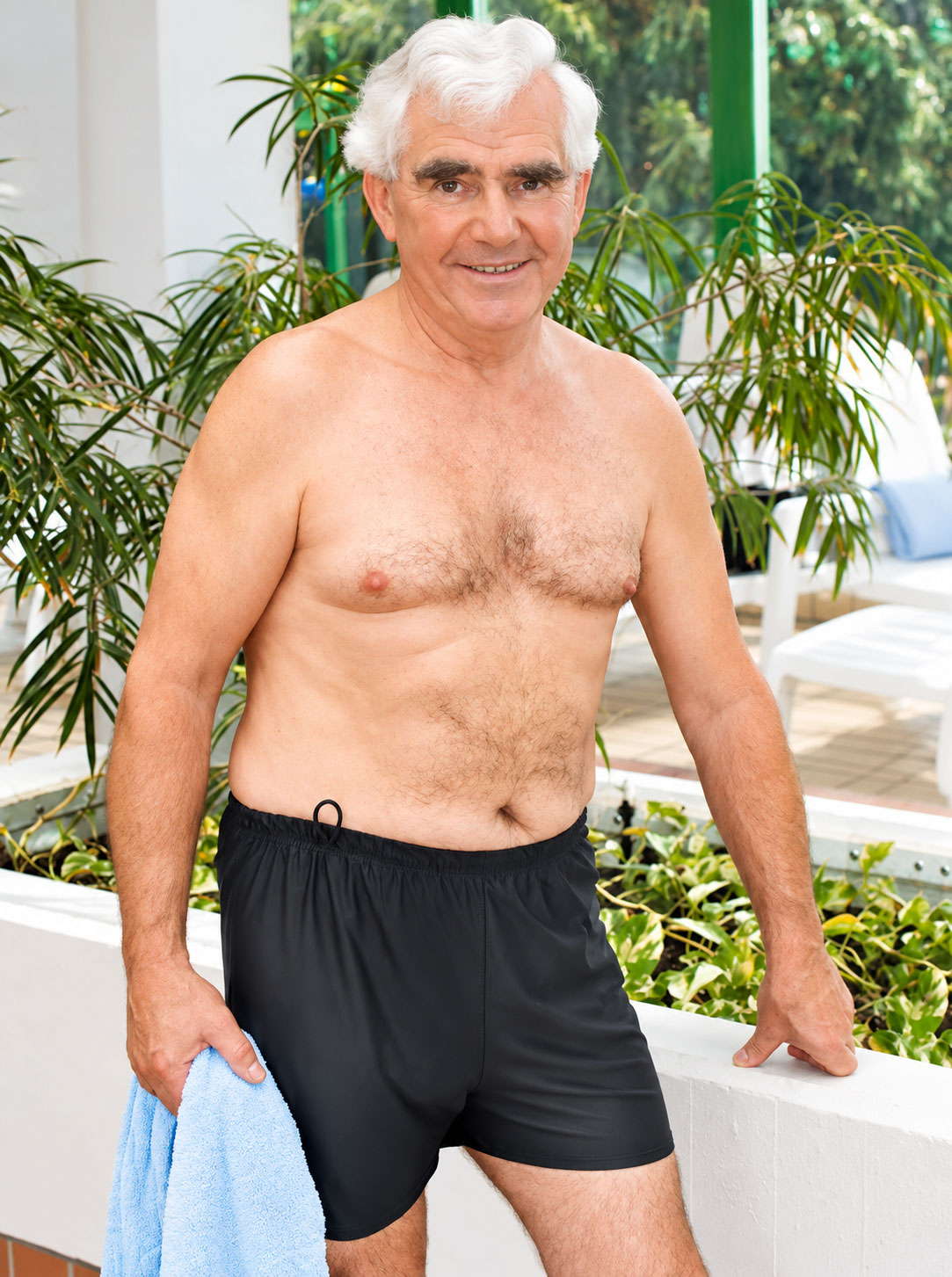 Ein älterer Mann mit grauen Haaren trägt schwarze Schwimmshorts und hält ein hellblaues Handtuch in der Hand. Er steht lächelnd vor einer grünen Pflanzenkulisse in einem hellen Innenbereich.