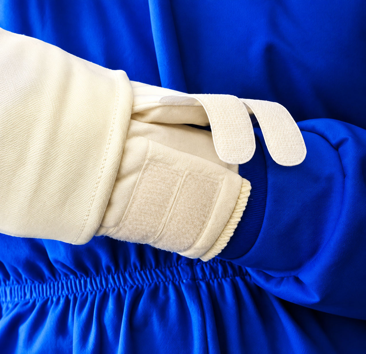 Beige Handschuh mit Klettverschluss, der einen blauen Kleidungsstoff berührt. Der Handschuh hat eine dicke, gepolsterte Textur. Im Hintergrund ist eine blaue, elastische Stoffstruktur sichtbar.