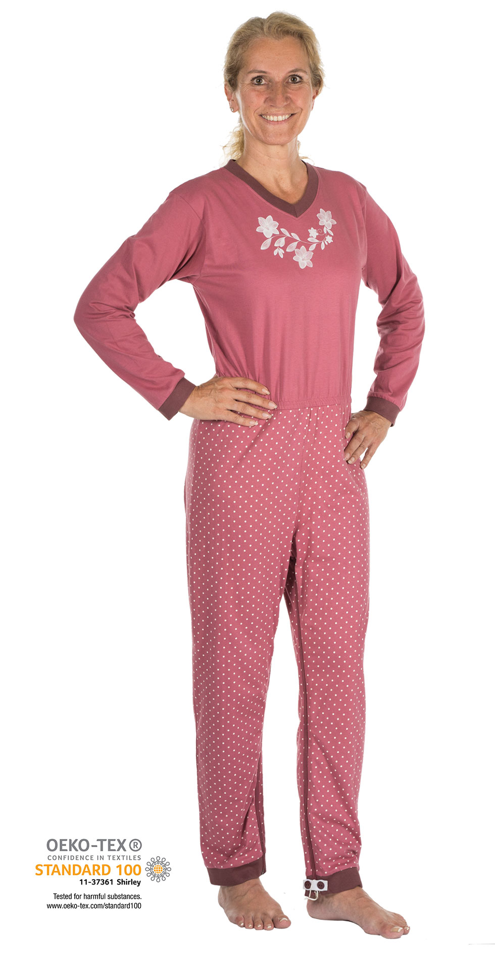 Ein Schlafanzug mit einem pinken Oberteil und gepunkteten Hosen wird von einer lächelnden Frau getragen. Der Schlafanzug hat eine Blumenstickerei. Der Hintergrund ist weiß. 

OEKOTEX®-Logo und Text sind im Bild zu sehen.