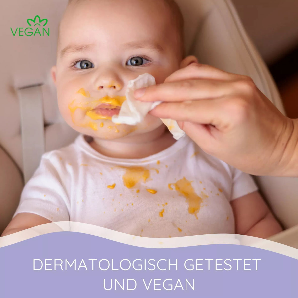 Ein Baby mit kleinen Essensflecken im Gesicht sitzt in einem Hochstuhl. Eine Hand wischt mit einem Tuch den Mund des Babys ab. Oben links steht das Wort -VEGAN-. Unten steht: -DERMATOLOGISCH GETESTET UND VEGAN-.
