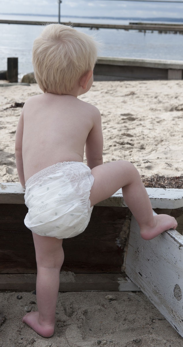 Ein kleines Kind mit blonden Haaren trägt eine Windel und versucht, über eine Holzbarriere zu klettern. Der Hintergrund zeigt einen Sandstrand und Wasser, mit einem Pier in der Ferne.