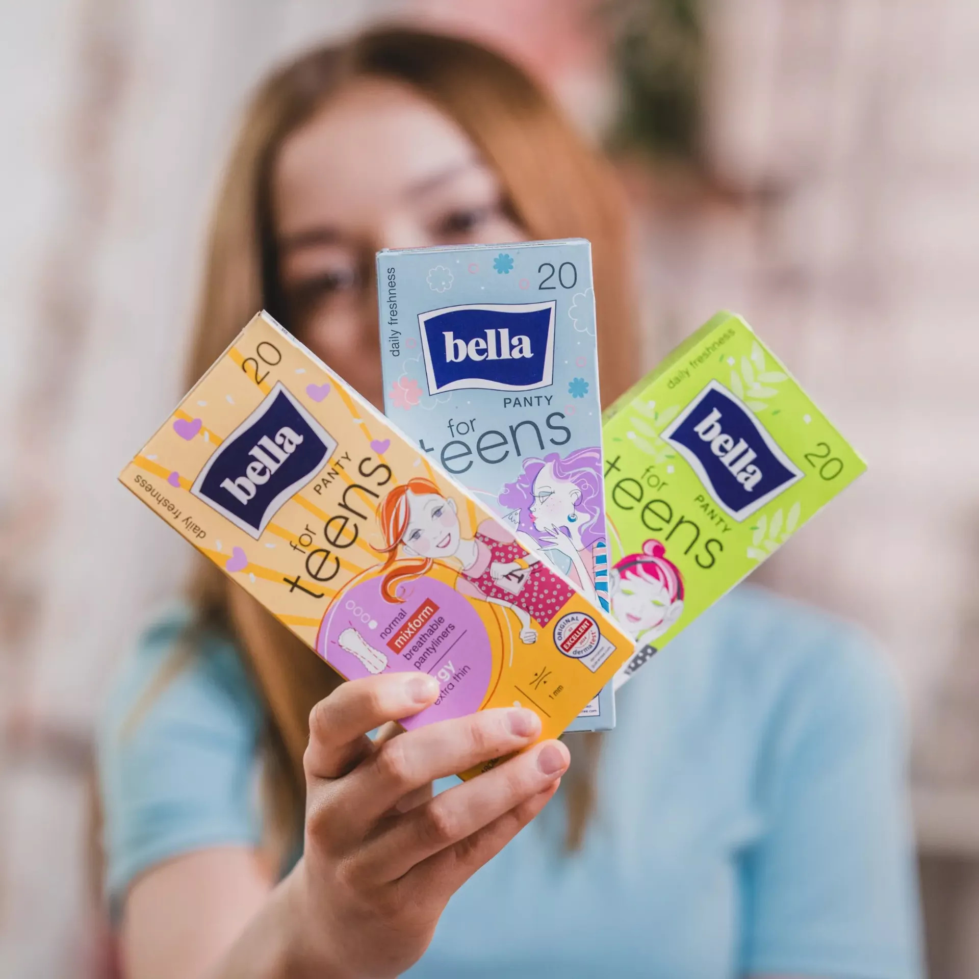 Drei Verpackungen von -bella for teens- Damenbinden werden von einer Person gehalten. Die Packungen sind in verschiedenen Farben und Designs gestaltet und zeigen auf der Vorderseite Illustrationen, die auf die Zielgruppe hinweisen. Hintergrund ist unschar