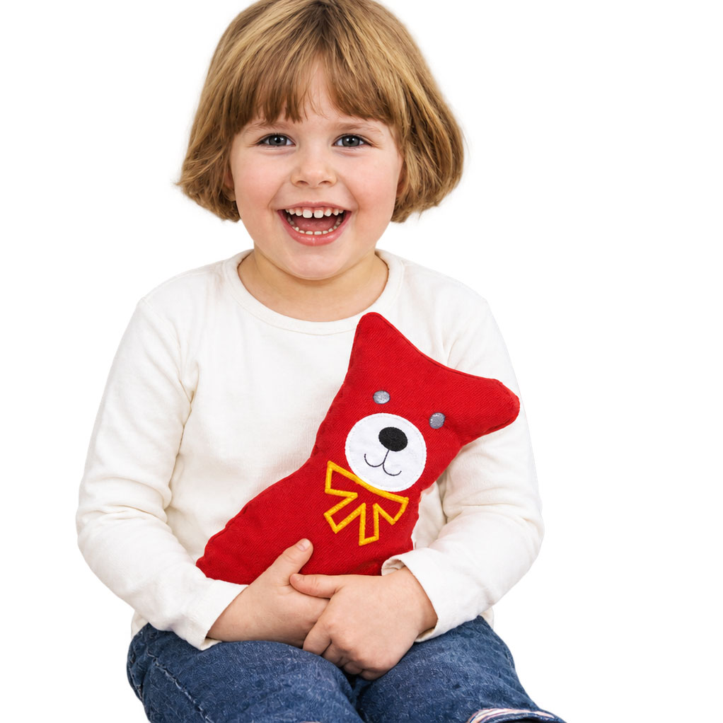 Ein lächelndes Kind hält ein rotes, bärenförmiges Stofftier mit einem weißen Gesicht und einer gelben Schleife in den Händen. Es sitzt vor einem hellen, einfarbigen Hintergrund.