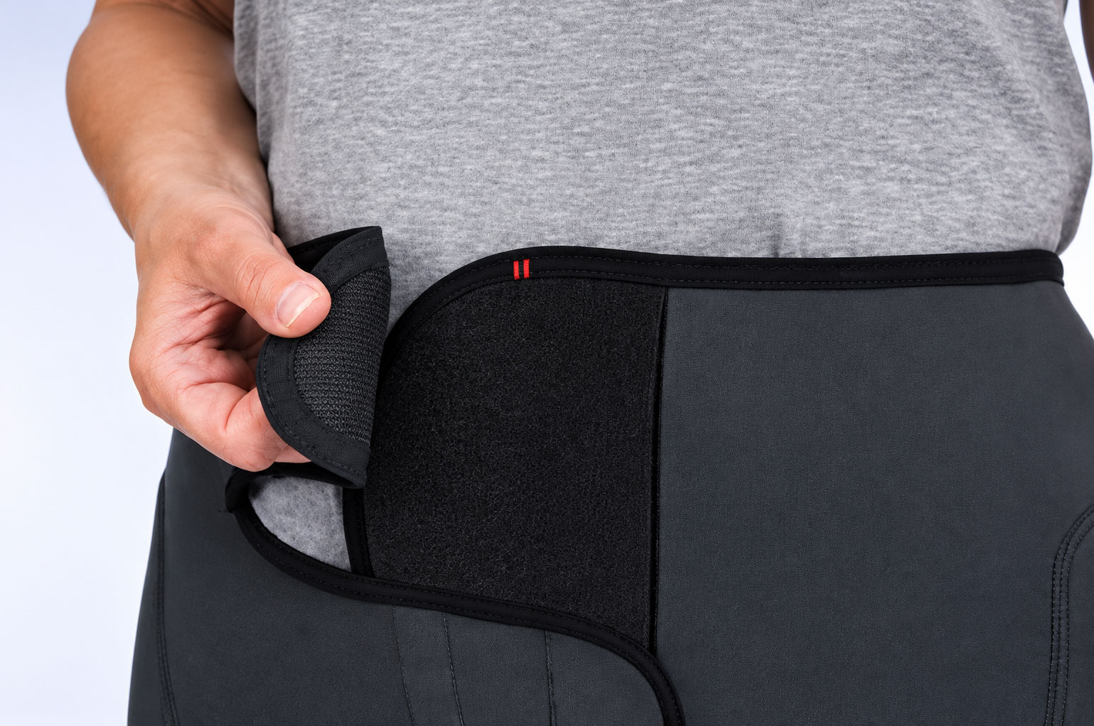 Eine Hand hält eine Klettverschlusstasche an einer schwarzen Inkontinenzhose, die an einem grau trikotierten Oberteil getragen wird. Der Hintergrund ist hell und neutral.